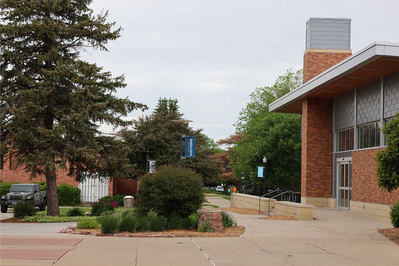 An image of Dakota State University Campus with a tree on the left, a bush in the center, and the Trojan Center on the right.