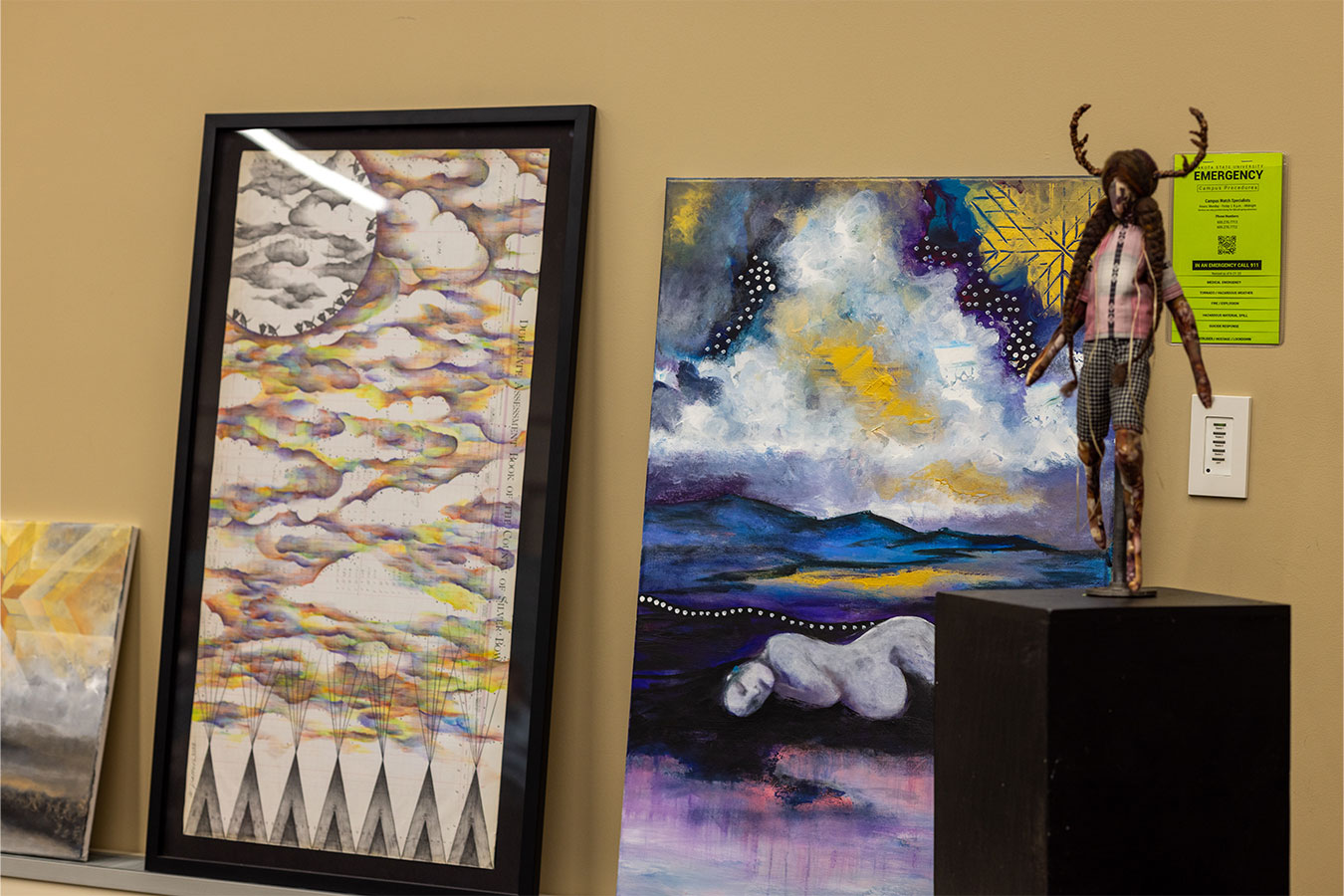 Pictured (l-to-r): A ledger drawing by Wade Patton featuring colorful clouds, a full moon, and seven tipis; a painting by J.White featuring a pregnant woman laying on the ground; a doll featuring braids and horns. 