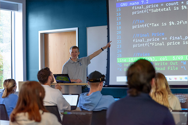 Computer science professor teaching a class in front of a screen displaying computer code.