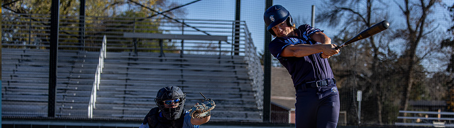 A batter swings hard at a ball as the catcher squats behind him. Both are wearing Trojan uniforms.