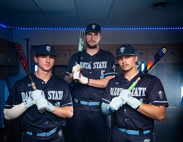 Three DSU baseball players pose with bats resting on their shoulders inside a locker room.