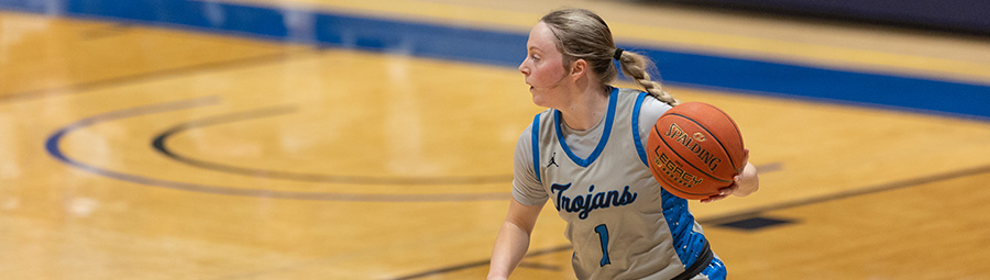 A young woman in a Trojans basketball jersey dribbles a basketball on an empty court.