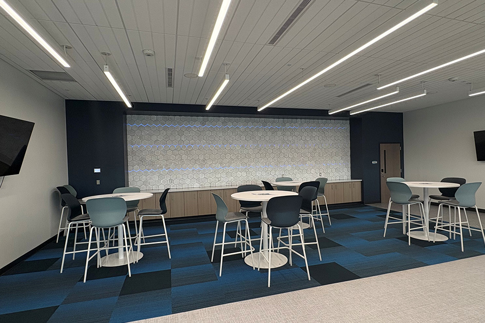 A large brightly lit room with high round tables placed spaciously on a patterend blue carpet. The far wall is covered in a hexagon pattern with blue neon lights.