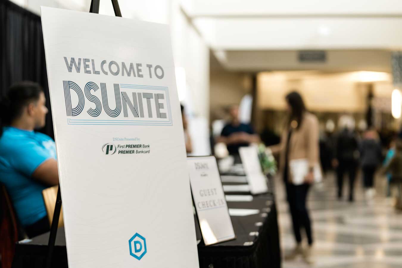 Photo of a sign reading Welcome to DSUnite presented by First PPREMIER Bank PREMIER Bankcard with an out of focus woman in the background. 