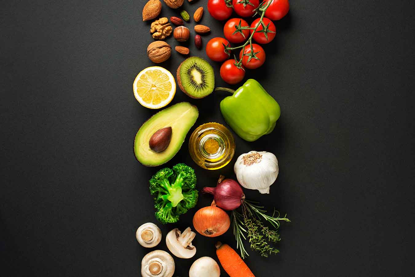 A grey background with fresh groceries placed in a line down the center featuring nuts, tomatoes, a lemon, kiwi, green pepper, avocado, jar, broccoli, garlic, onions, mushrooms, and a carrot. 