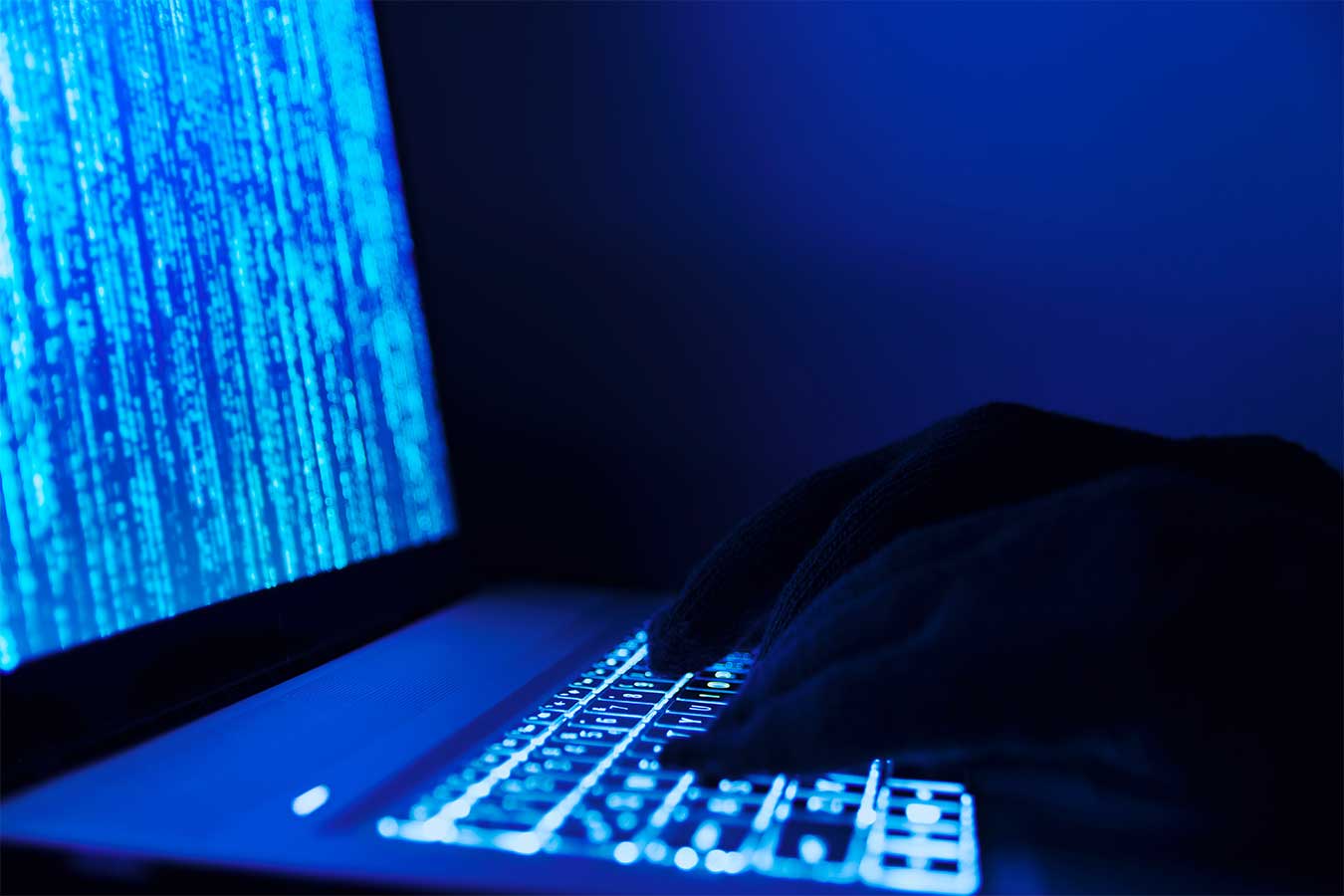A stock image of black gloved hands typing on a laptop with blue light emanating from it.  