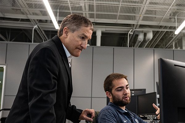 Student sitting at computer while an instructor wearing a suit looks over his shoulder. 