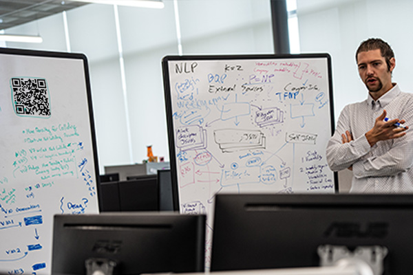Professor gesturing in front of a whiteboard filled with charts and text about coding structures