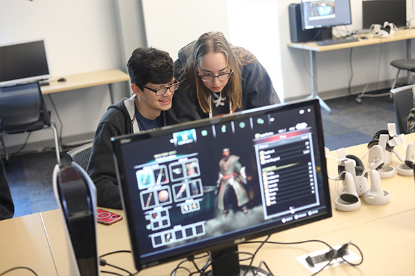 Computer Game Design students looking at a monitor. A playstation 5 and a monitor displaying a video game menu sits on the table.