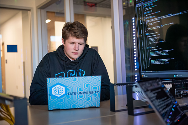 Computer science minor working on a laptop in front of a large screen displaying computer code.