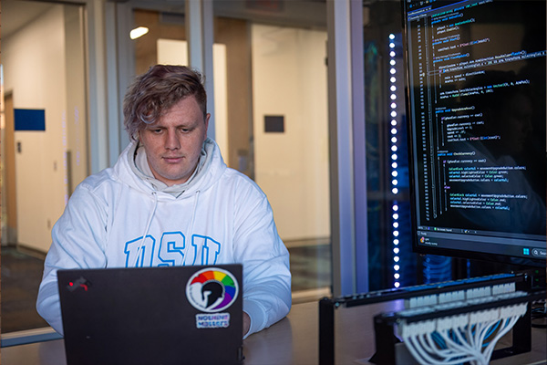 data privacy student working on a laptop near a large screen displaying code.