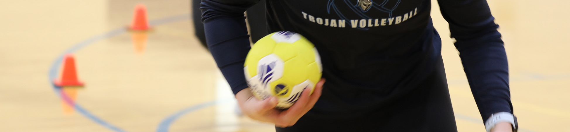 A Trojan student holding a handball, or maybe a small volleyball, in hand as she runs througha gymnasium aroundn orange cones.