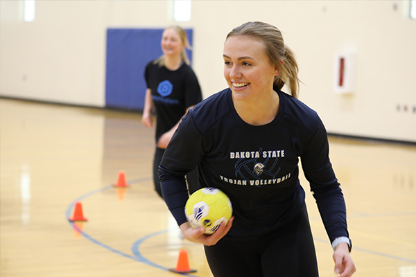 A Trojan student holding a handball, or maybe a small volleyball, in hand as she runs througha gymnasium aroundn orange cones.
