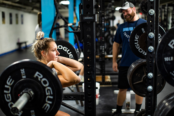A DSU student squatds underneath heavy weights in the gym, coached by a strength and conditioning instructor.