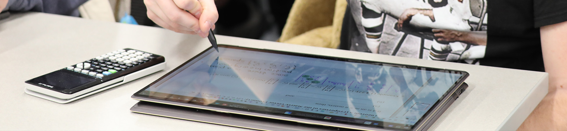 A student writing math equations on a tablet computer with a graphic calculator nearby on the desk.
