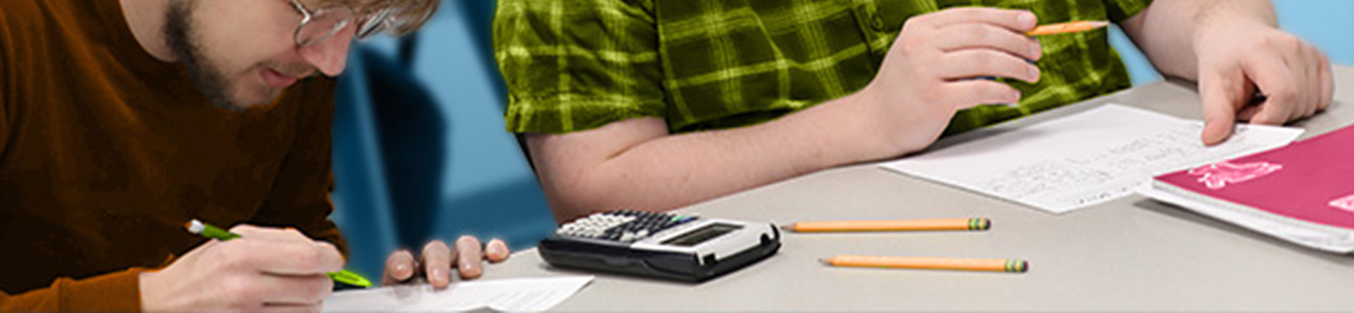 Two math students working on equations with pencil and paper. A graphic calculator and extra pencils are on the desktop.