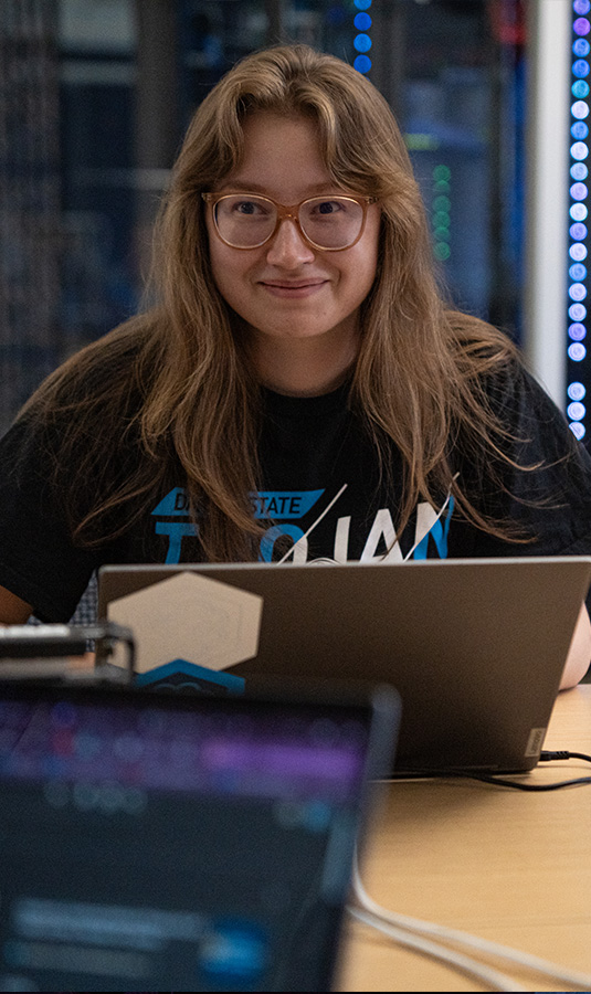 Cyber operations masters student smiling in front of a glowing bank of servers.